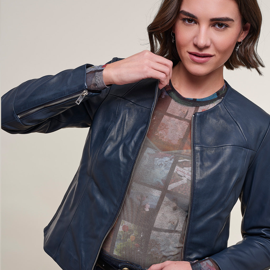 A woman wearing a dark navy blue faux leather jacket over a sheer patterned top, posing against a neutral beige background.