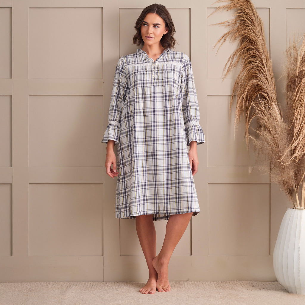 A woman stands barefoot in front of a beige paneled wall wearing a long-sleeve plaid nightshirt with a shawl collar. The nightshirt has a white base with gray and black plaid lines.