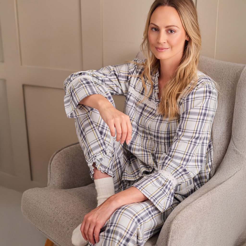 A woman wearing a long-sleeve plaid pyjama top in cream, white, and navy blue, sitting a grey chair against a beige paneled wall.