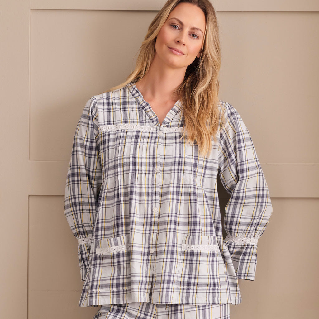 A woman wearing a long-sleeve plaid pyjama top in cream, white, and navy blue, standing against a beige paneled wall.