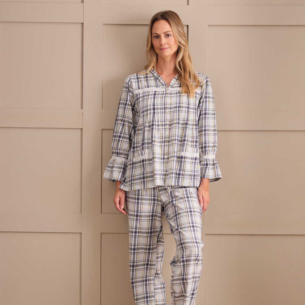 A woman wearing a long-sleeve plaid pyjama top in cream, white, and navy blue, standing against a beige paneled wall.