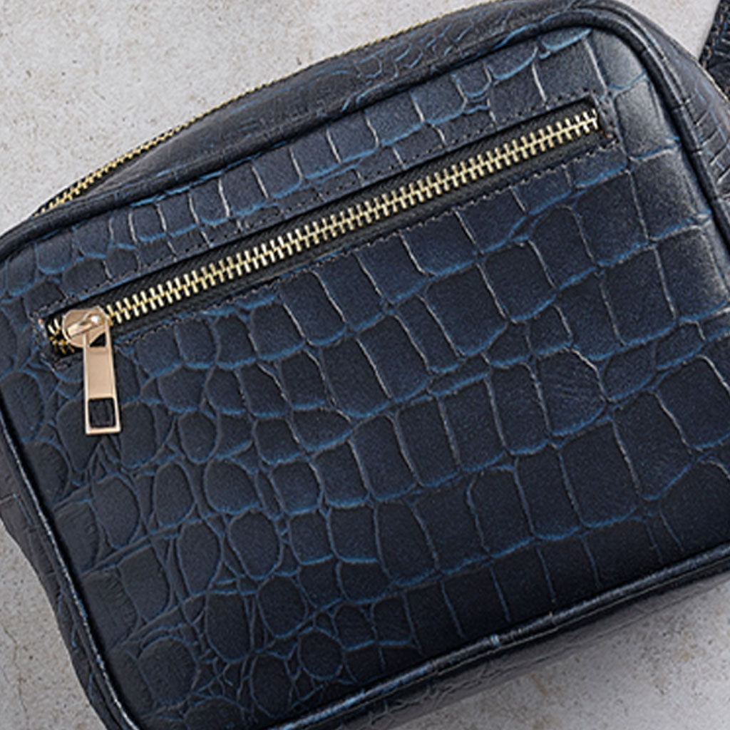 A close-up of a dark blue crocodile-embossed leather bag with a gold zipper.