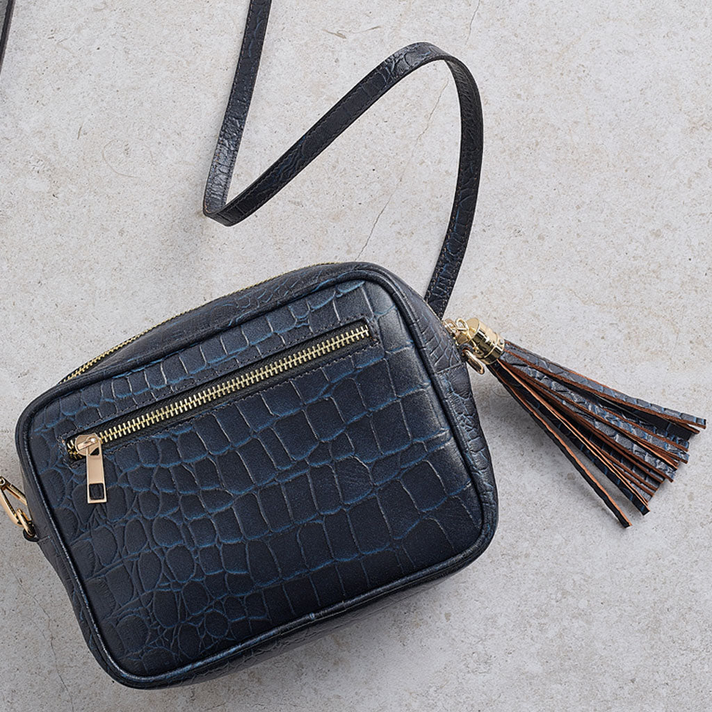 A dark blue crocodile-embossed faux leather crossbody bag with a gold zipper, a matching shoulder strap, and a brown tassel charm with gold hardware, displayed on a light gray textured background.