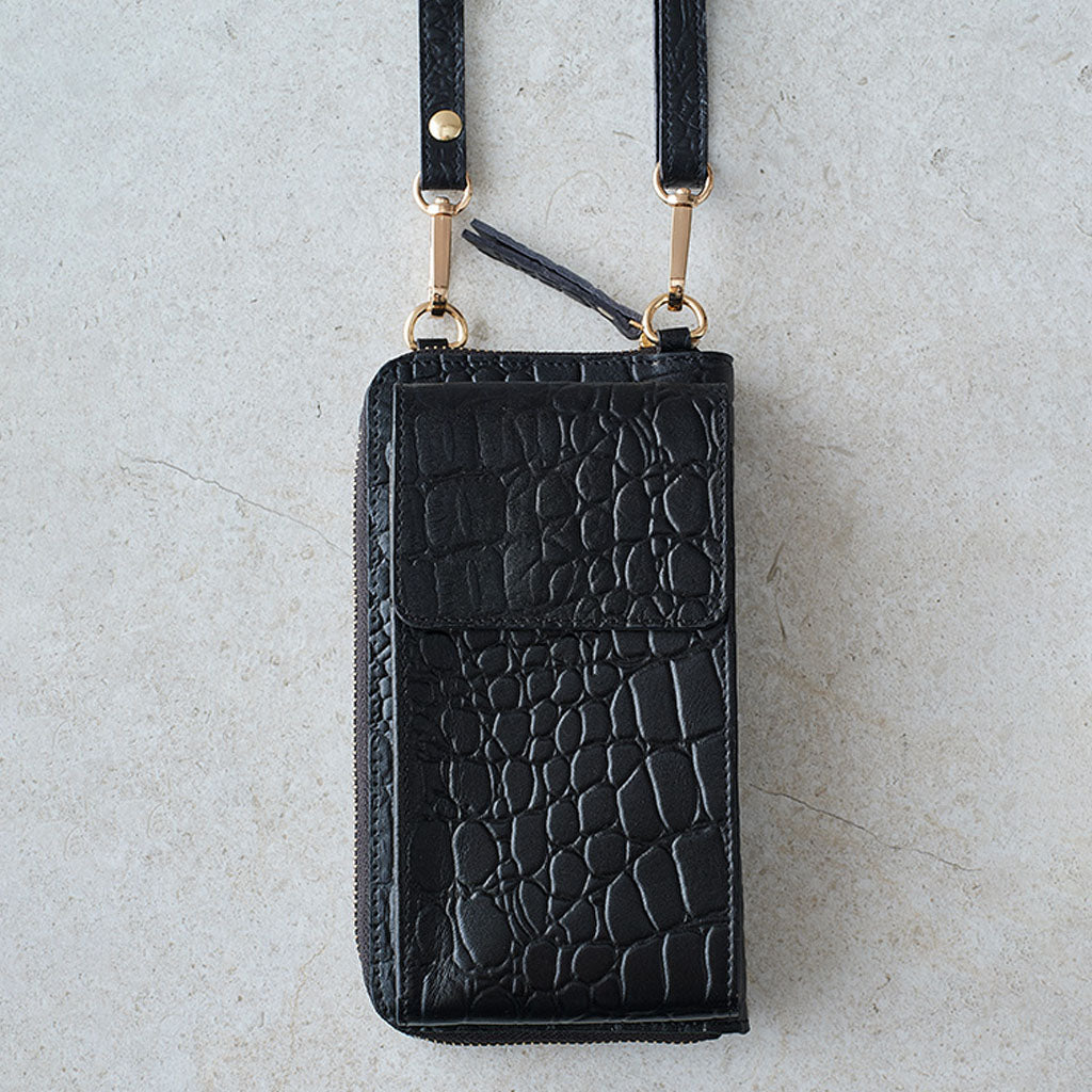 Black crocodile-embossed leather zip-around phone wallet with a matching shoulder strap and gold-tone hardware, shown flat on a light textured surface.