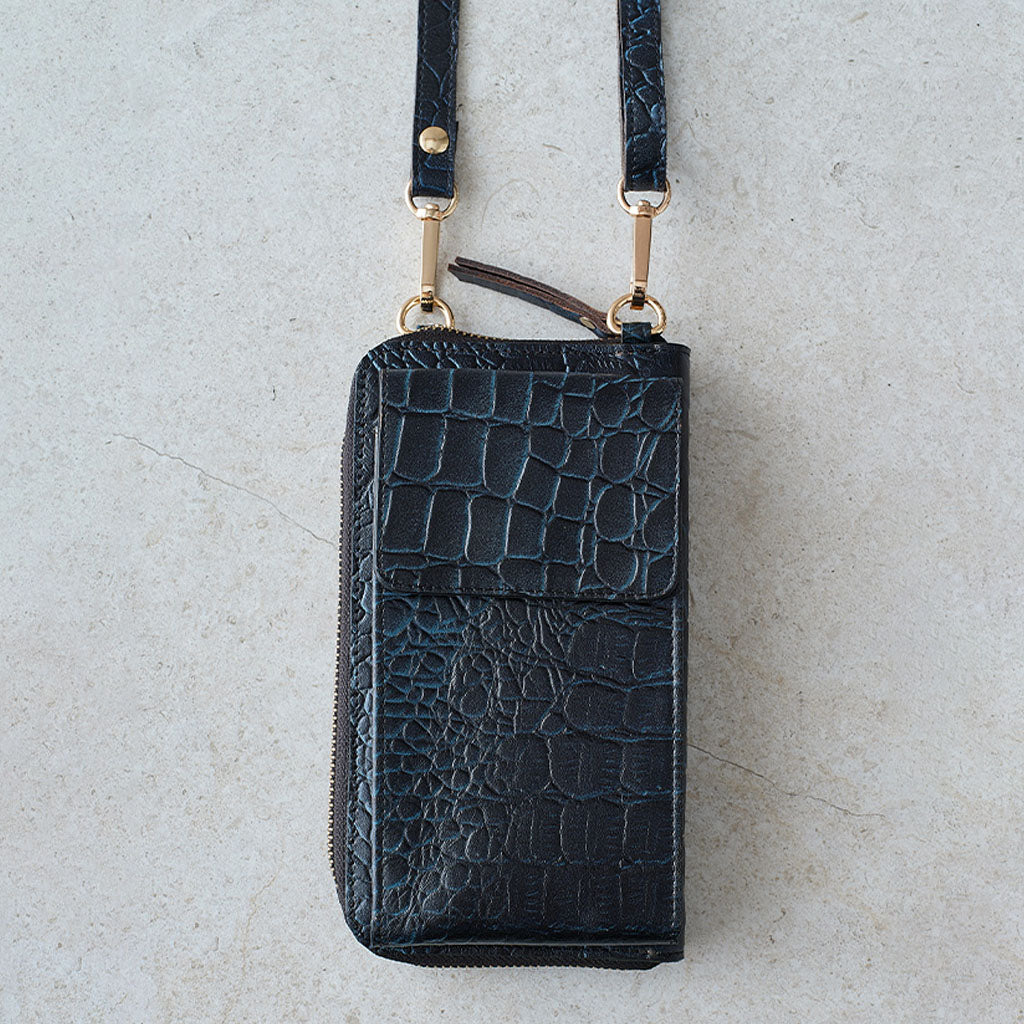 A black crocodile-embossed leather zip-around phone wallet with a matching wrist strap and gold-tone hardware, laid flat on a light textured surface.