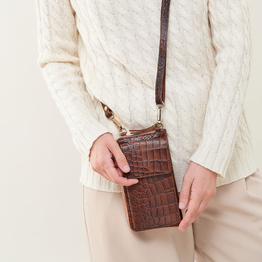 A person wearing a cream cable-knit sweater and beige pants holds a brown crocodile-embossed leather crossbody phone bag with a matching strap against a light beige background.