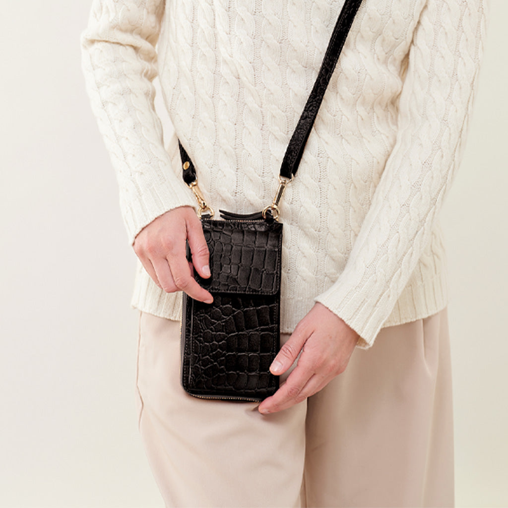 A person wearing a cream cable-knit sweater and beige pants holds a small black crocodile-embossed crossbody bag with a gold-tone clasp and strap.