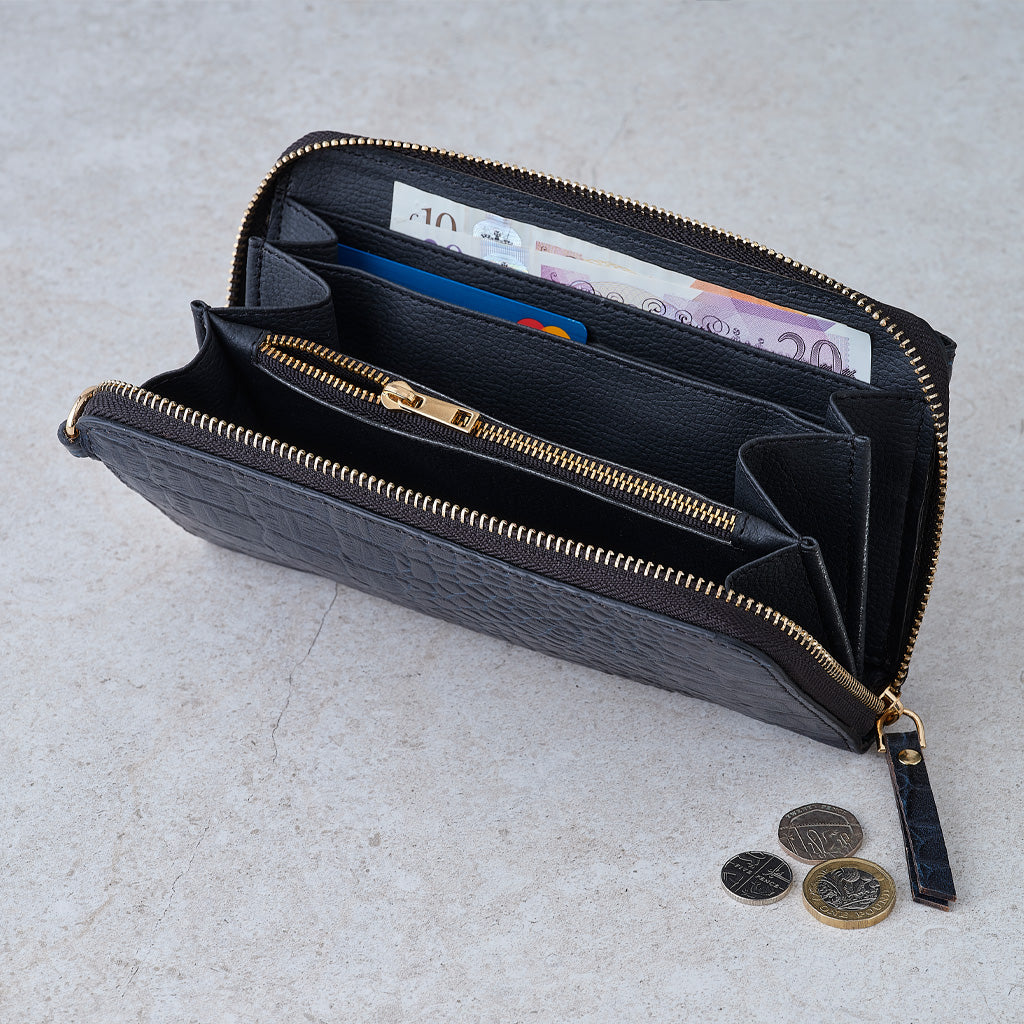 A black embossed leather zip-around wallet shown open, revealing multiple interior compartments, card slots, and a gold-tone zipper. Euro banknotes and coins are placed inside and beside the wallet on a light stone surface.