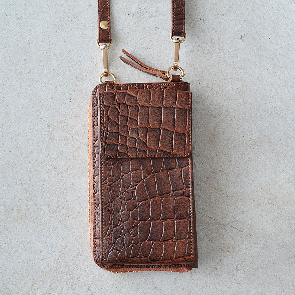 A brown crocodile-embossed leather phone wallet with a matching shoulder strap and gold-tone hardware, laid flat on a light gray textured surface.
