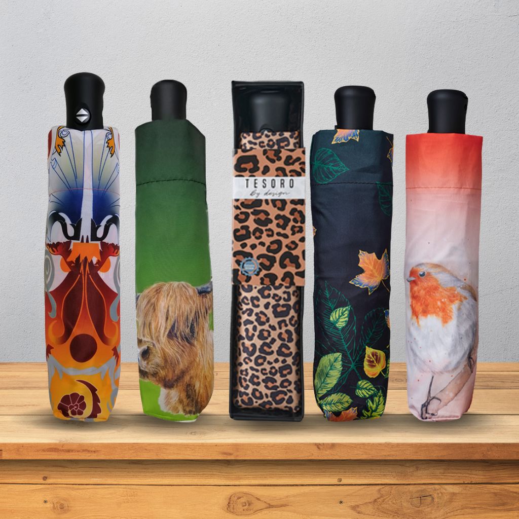 Five folded umbrellas with black handles are lined up on a wooden shelf against a light gray wall. Each umbrella cover has a different printed design: a colorful abstract floral pattern, a green background with a highland cow portrait, a leopard print with a small bird, a dark green and orange leaf pattern, and a pink-to-orange gradient with a bird.