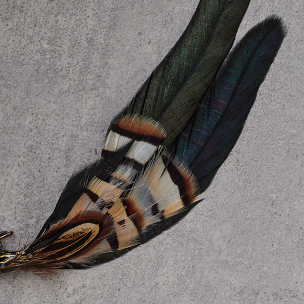 A decorative feather brooch featuring two long French Partridge feathers with black, brown, and cream bands, attached to an ornate antique bronze safety pin, displayed on a textured gray background.