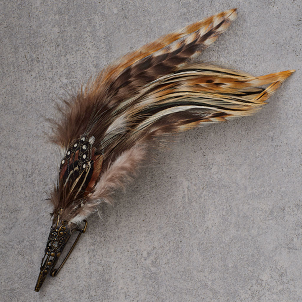 A decorative feather hair clip with a long brown, black, and tan patterned feather attached to a dark metal base, shown on a textured gray background.