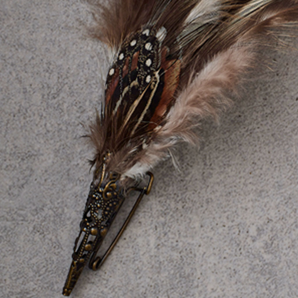A decorative feather pin with brown and beige feathers, a black and white patterned center, and an ornate antique bronze metal base with a safety pin clasp, shown on a textured gray background.