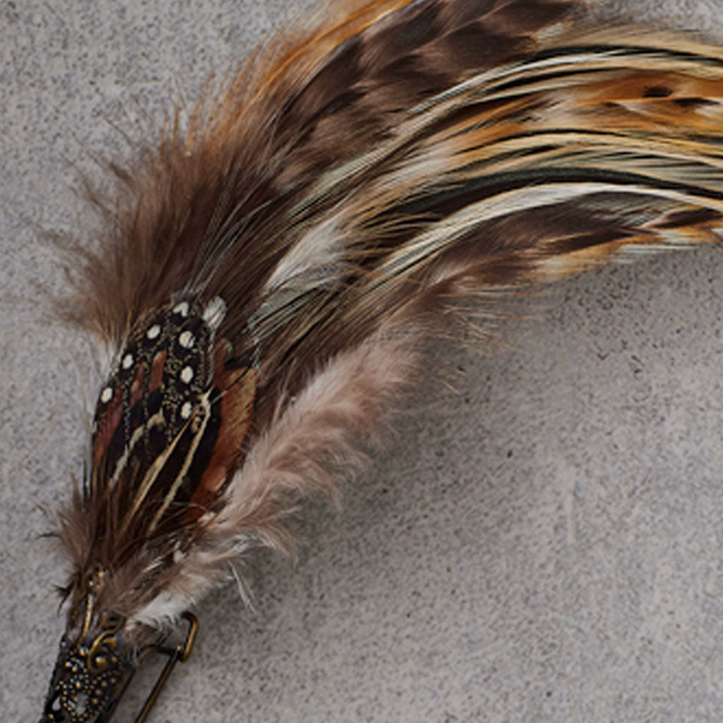 A decorative feather brooch featuring two long Spotted Guinea Fowl feathers, brown, and cream bands, attached to an ornate antique bronze safety pin, displayed on a textured gray background.