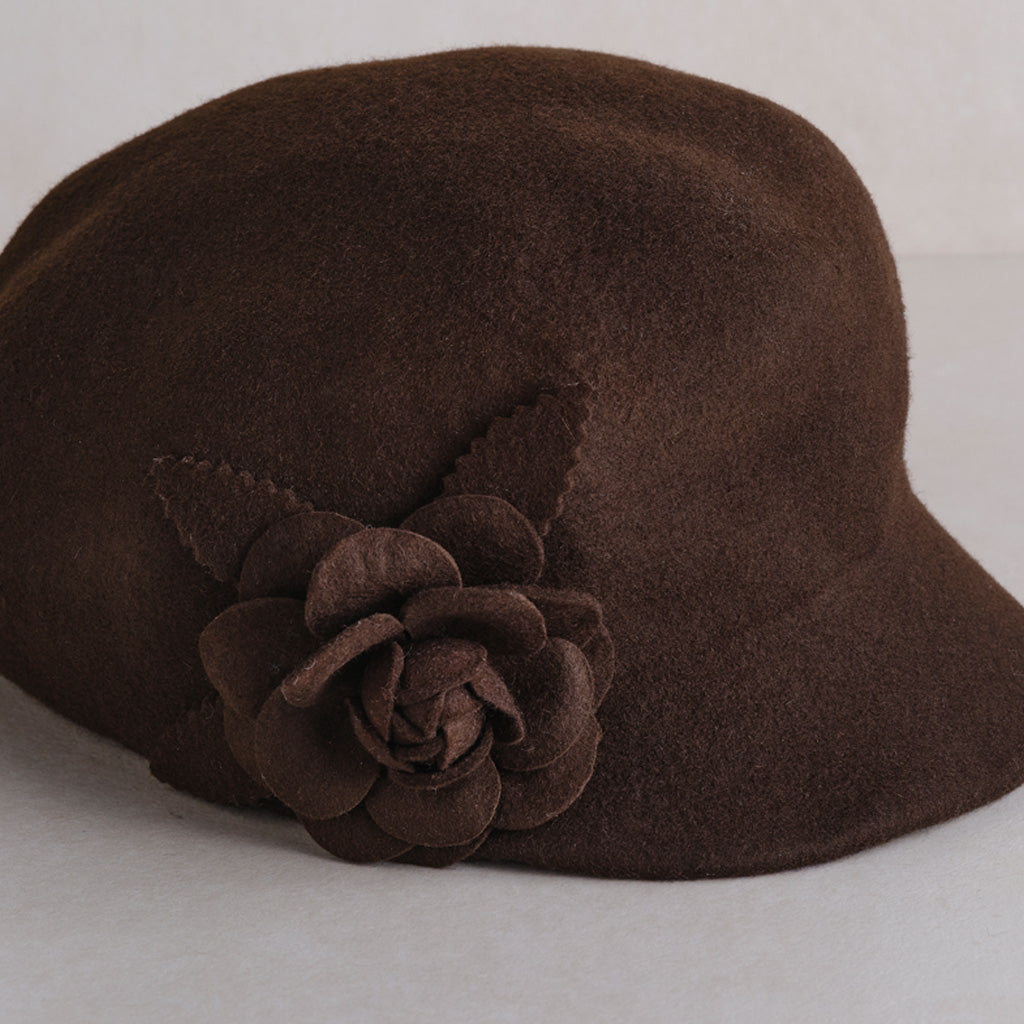 A dark brown felt cloche hat featuring a matching fabric flower and leaf embellishment on the side, set against a light neutral background.