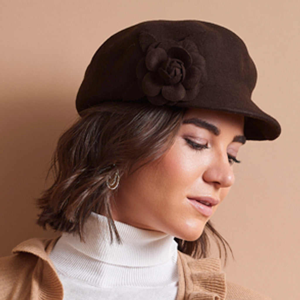 A woman wearing a dark brown felt-style newsboy cap with a large fabric rose embellishment on the side, paired with a white turtleneck and tan jacket against a plain beige background.