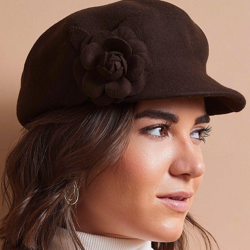 A close-up of a woman wearing a dark brown felt newsboy cap with a large fabric flower embellishment on the side, set against a solid beige background.