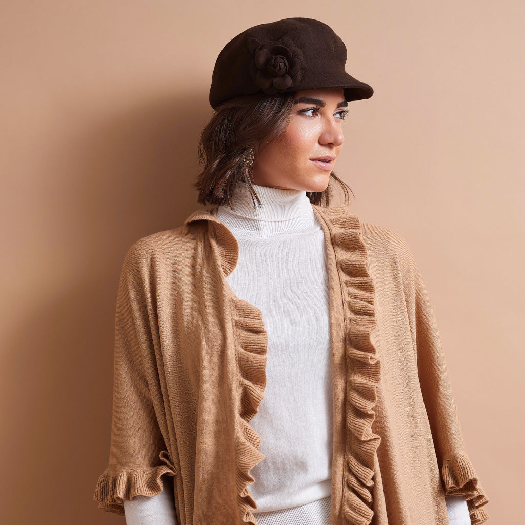 A woman wearing a brown felt newsboy cap with a fabric flower, a white ribbed turtleneck sweater, and a camel beige open-front cardigan with ruffle trim, posed against a solid warm beige background.