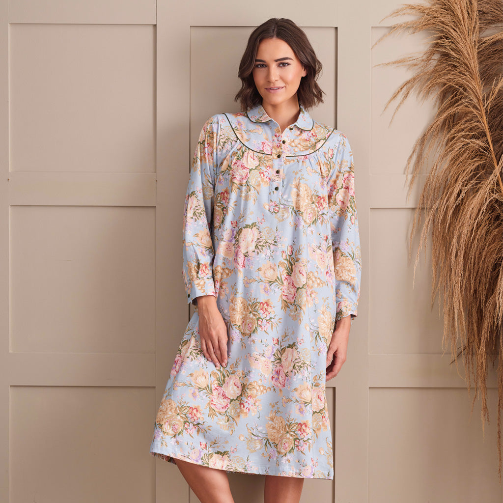 A woman models a light blue long-sleeve nightgown with a vintage floral print in pink, beige, and green, standing against a beige paneled wall with dried pampas grass on the side.