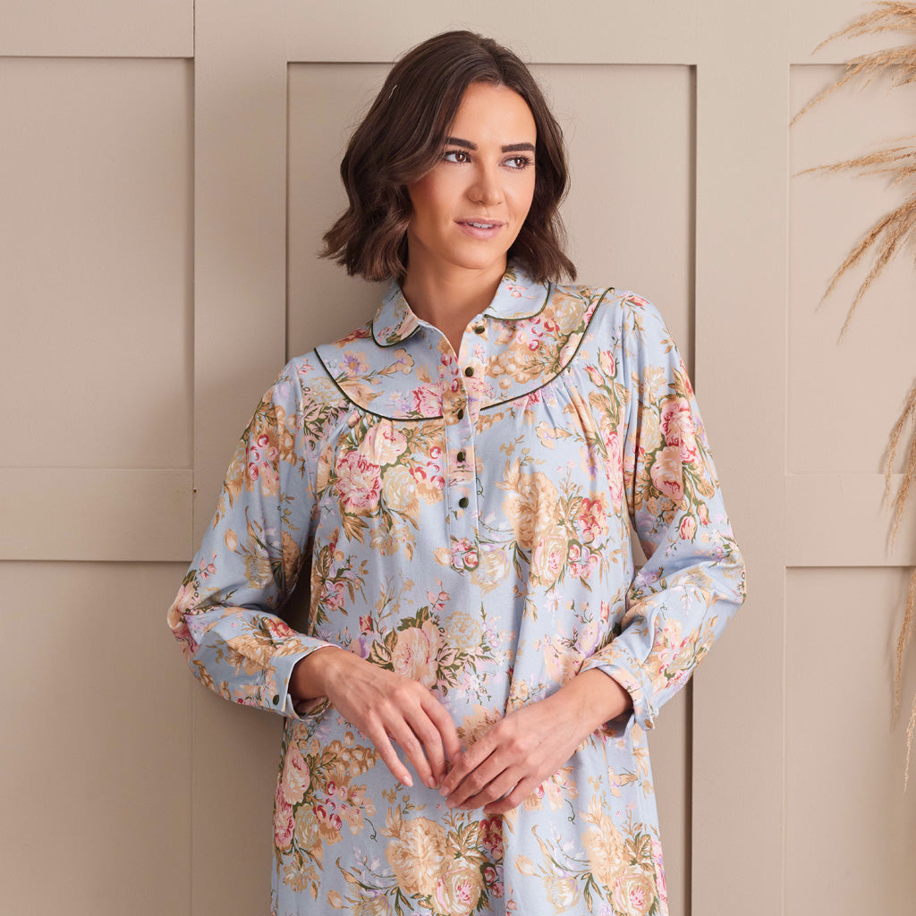 A woman wearing a light blue floral long-sleeve button-up shirt with a Peter Pan collar and black piping, standing against a beige paneled wall.