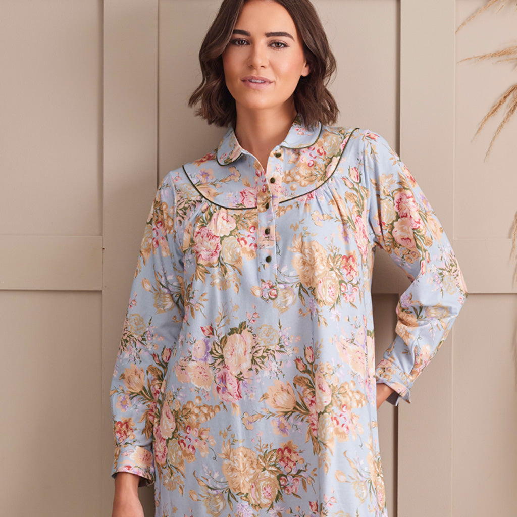 A woman wearing a light blue floral long-sleeve tunic shirt with a ruffled collar and button placket, standing against a beige paneled wall.