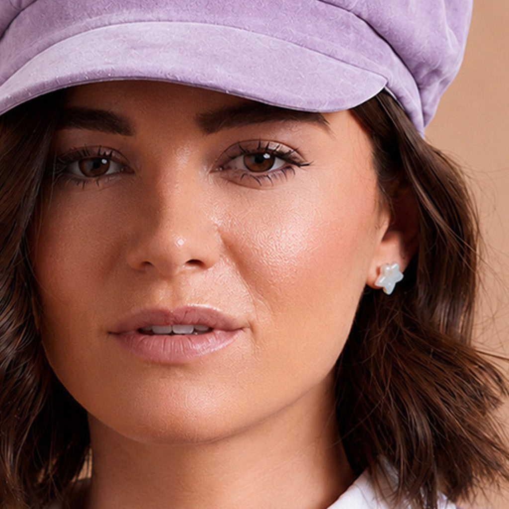 Close-up of a woman wearing a lavender velvet newsboy cap and a small pearl star-shaped stud earring, against a warm beige background.