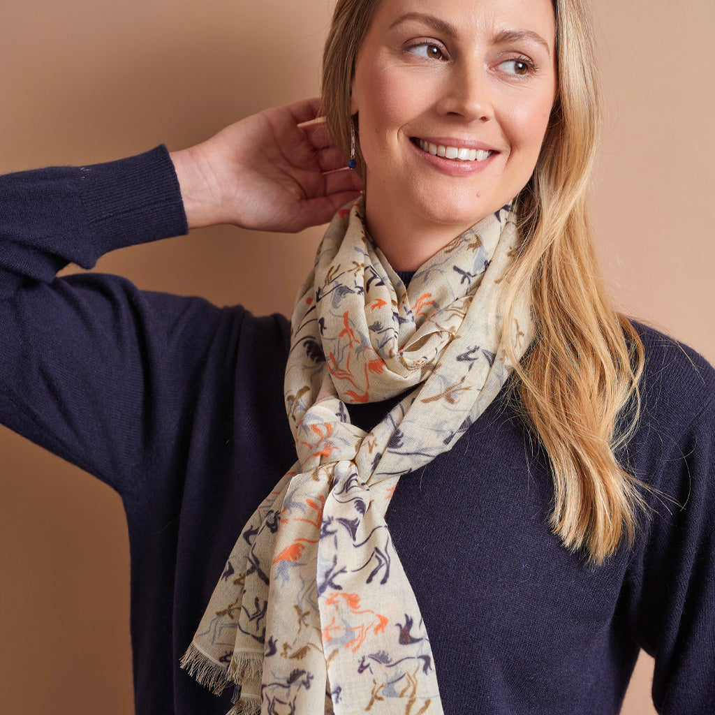 A woman wearing a navy blue knit sweater and a cream-colored scarf with a colorful animal print, posed against a solid warm beige background.