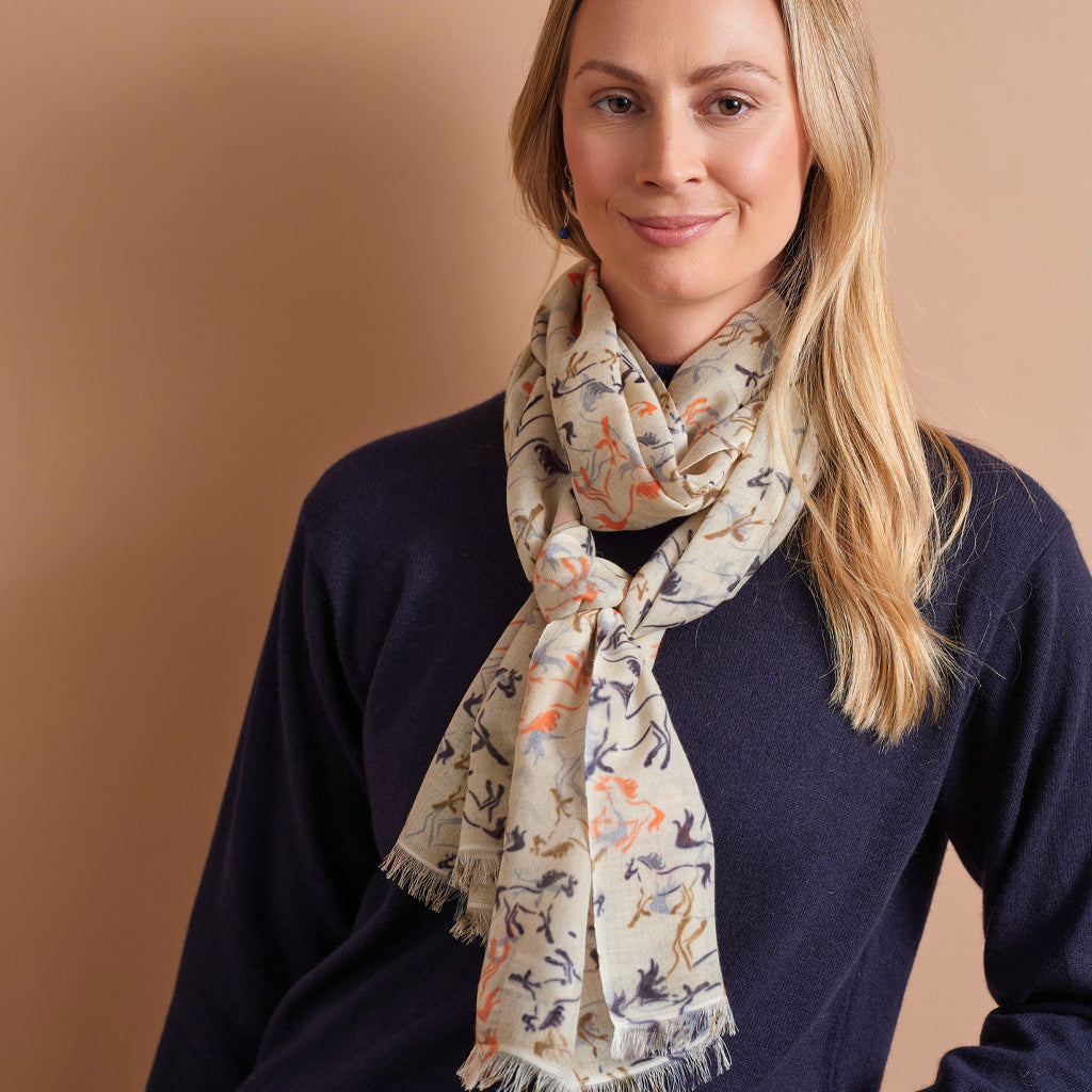 A woman wearing a navy blue sweater and a cream-colored scarf with a multicolor horse pattern, posed against a solid warm beige background.