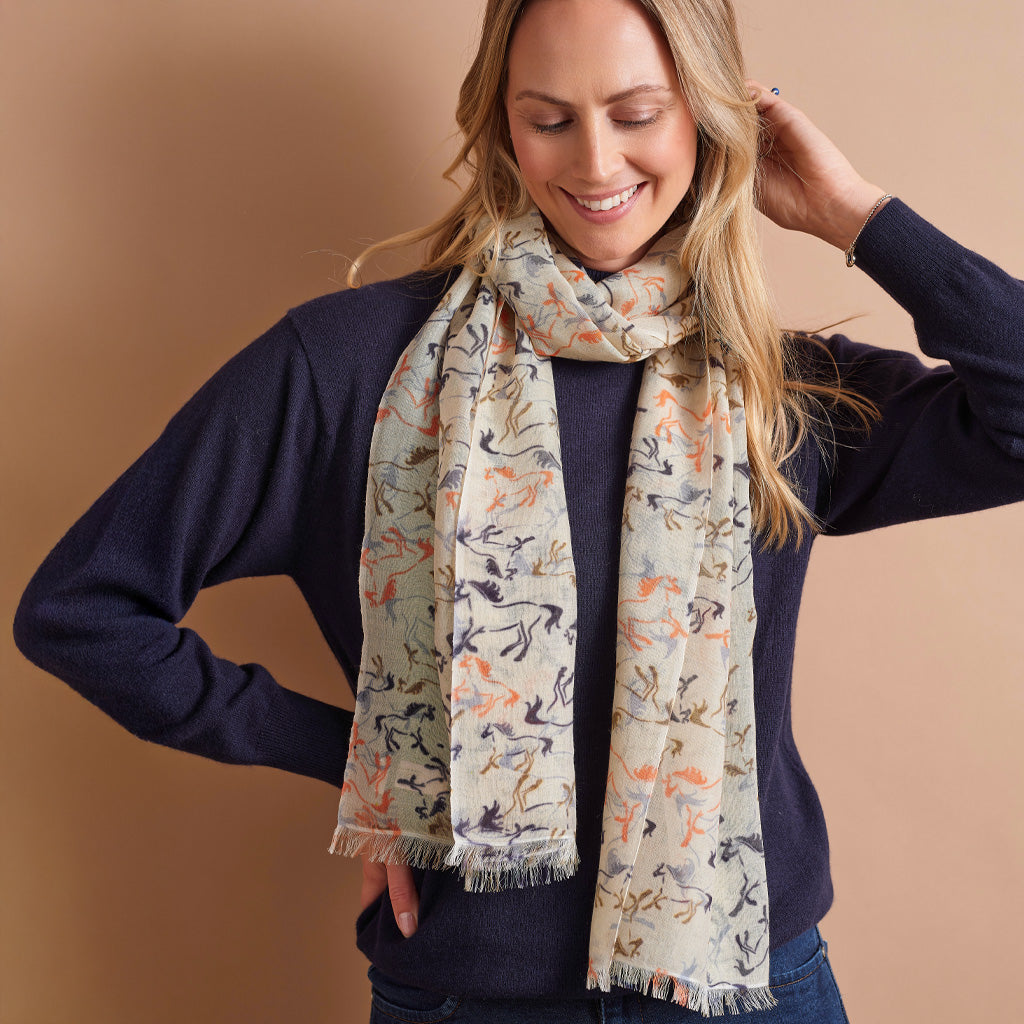 A woman wearing a navy blue sweater and jeans models a light beige scarf with a multicolor abstract pattern of birds and horses, set against a solid warm tan background.