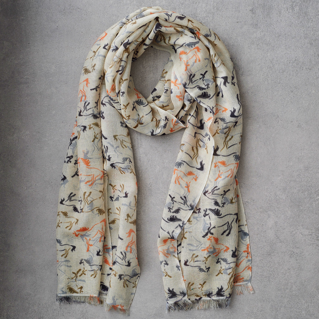 A lightweight cream scarf with an all-over abstract brushstroke pattern in orange, gray, and dark charcoal, styled in a loose loop on a textured gray background.