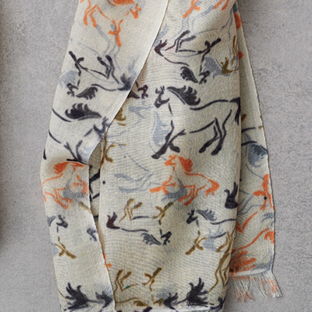 A cream-colored scarf with an abstract brushstroke pattern in black, orange, and muted blue, draped against a textured gray background.