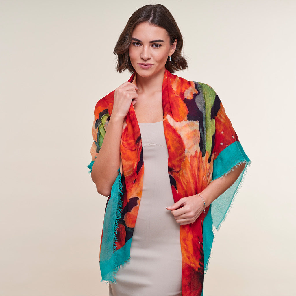 A woman modeling a lightweight, multicolored abstract print shawl with fringed edges over a light gray dress, set against a plain beige studio background.