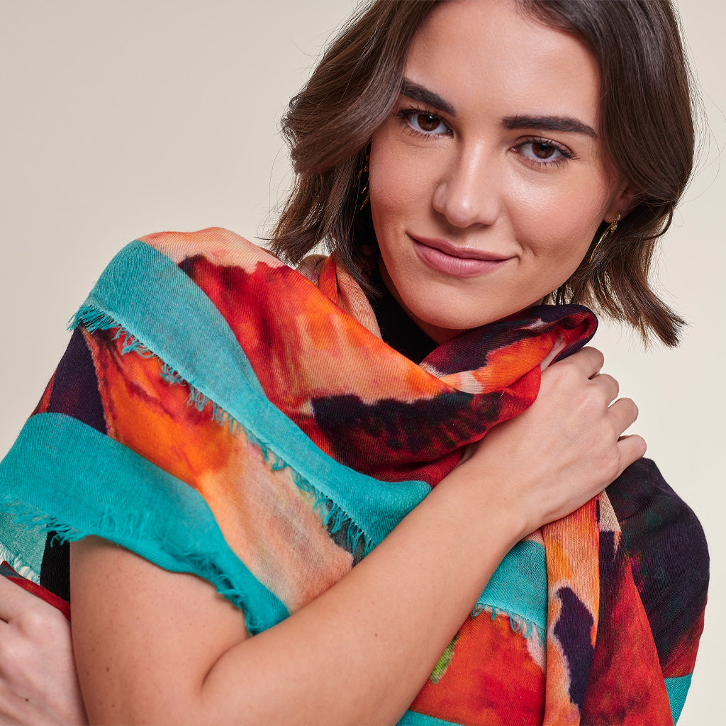 A woman wearing a lightweight, multicolored abstract print scarf with bright turquoise, orange, red, and black tones, photographed against a solid light beige background.
