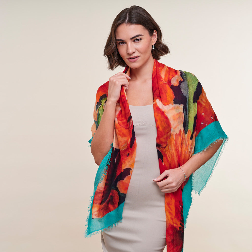 A woman modeling a lightweight, semi-sheer scarf with a vibrant abstract floral print in orange, red, green, and turquoise, worn over a light beige dress against a plain beige background.