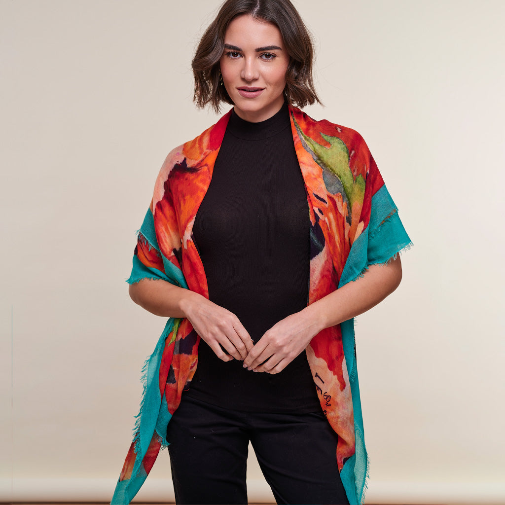 A woman modeling a vibrant abstract poppy print shawl with red, orange, teal, and green tones over a black ribbed turtleneck and black pants, set against a plain beige studio background.