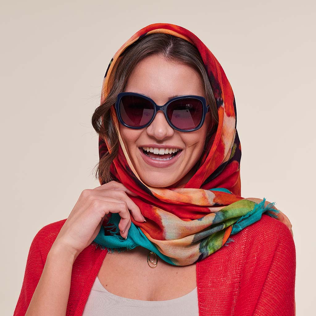 A smiling woman wearing a vibrant multicolor scarf with red, orange, teal, and green tones, styled with dark sunglasses and a red cardigan against a solid beige background.