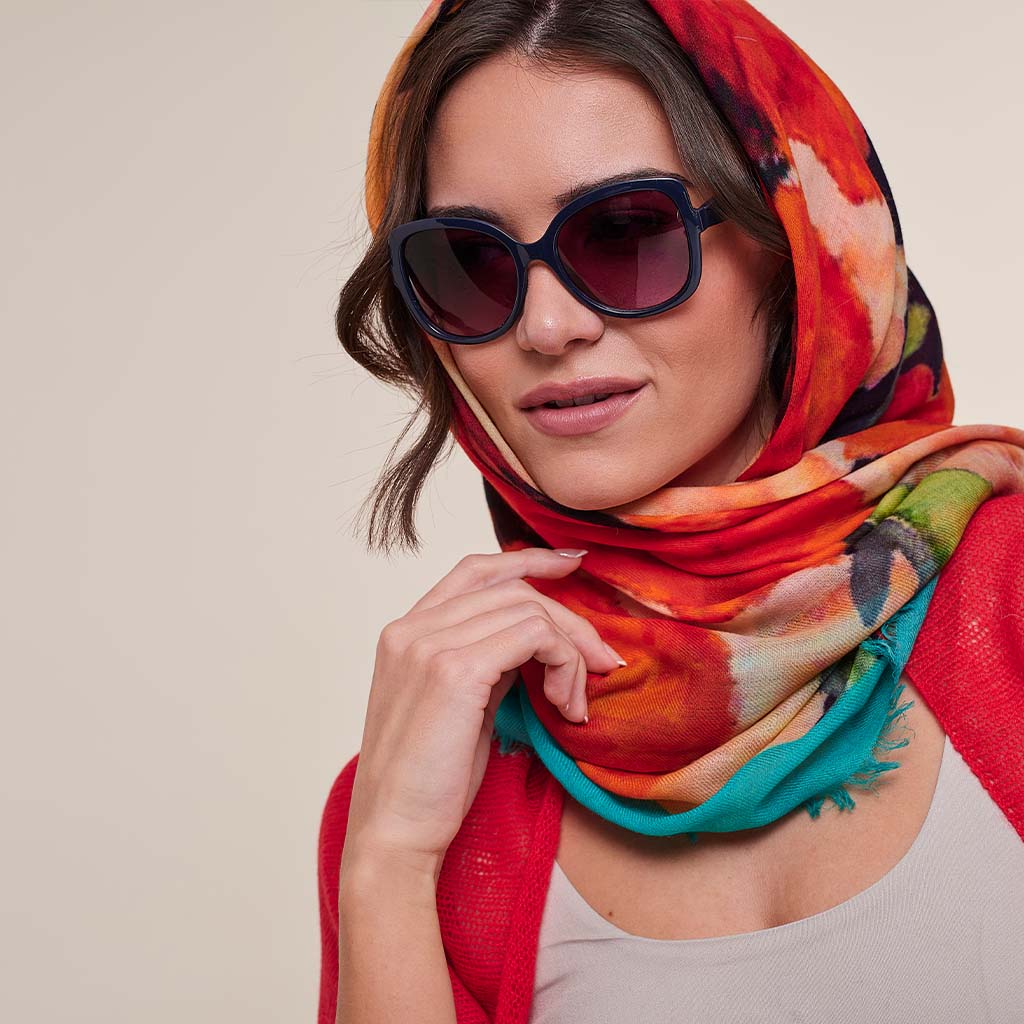 A woman wearing a vibrant, multicolored abstract print scarf in shades of red, orange, teal, and green, styled with dark sunglasses and a red cardigan against a neutral beige background.