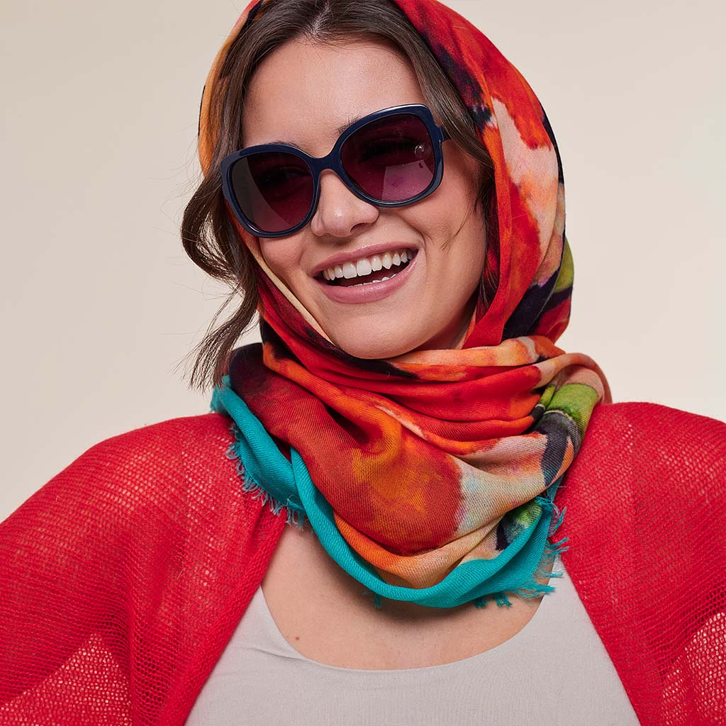 A smiling woman wearing a vibrant multicolor abstract print scarf in shades of orange, red, teal, and black, styled with dark sunglasses and a red open-front cardigan.