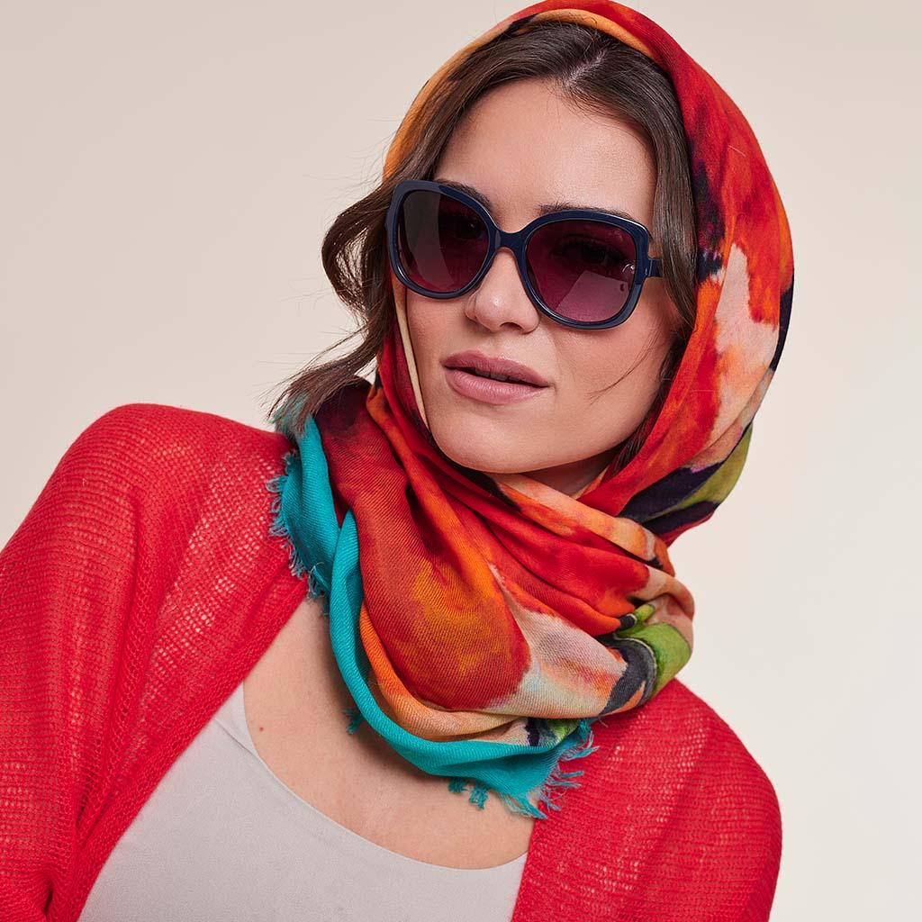 A woman wearing a vibrant, multicolored abstract-print headscarf and a red knit cardigan, styled with dark sunglasses against a plain light beige background.