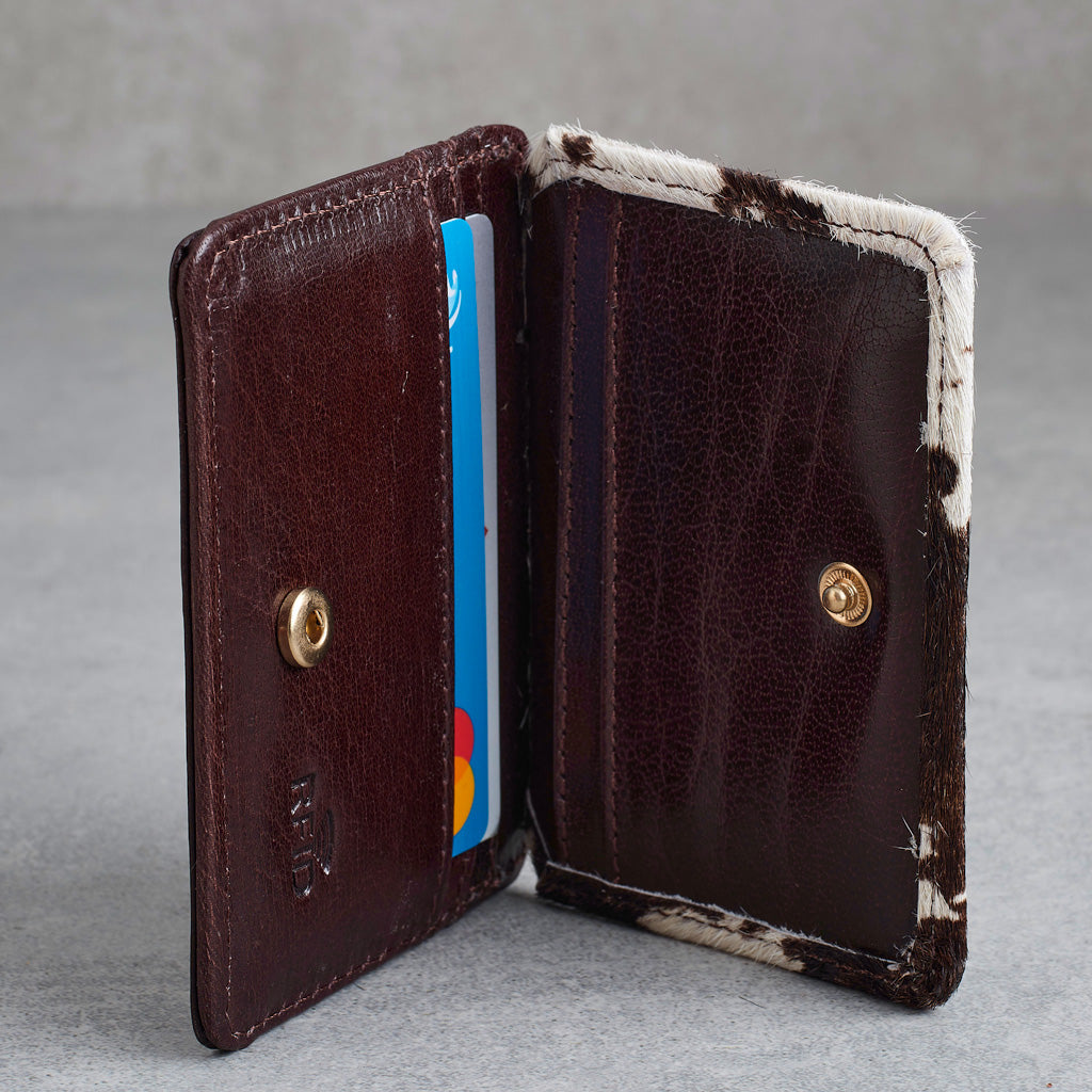 A dark brown leather wallet shown open, featuring a cowhide-textured trim with white and black patches, a gold snap button closure, and card slots with a blue and multicolor card visible inside, set against a neutral gray background.