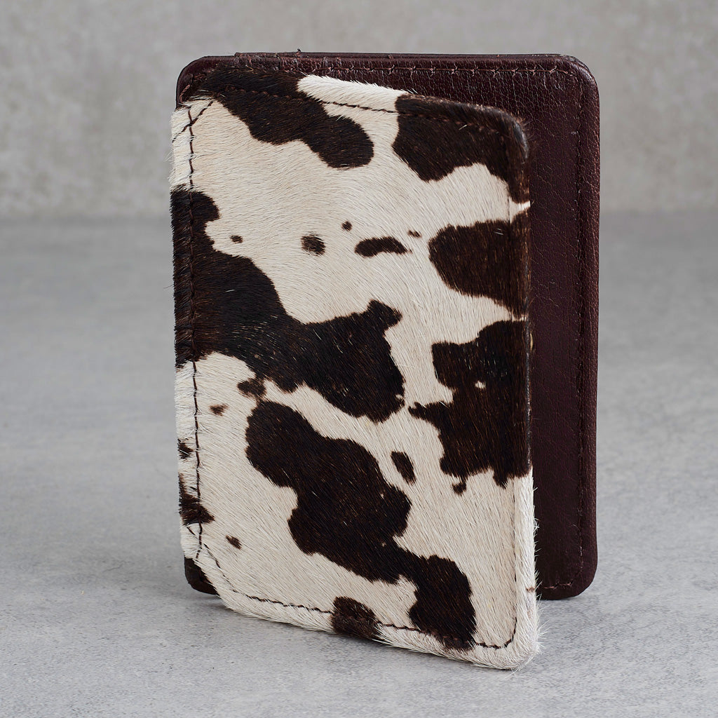 A small leather card holder or passport cover with a brown and white cowhide pattern on the front and a solid dark brown leather back, shown standing upright on a gray surface.