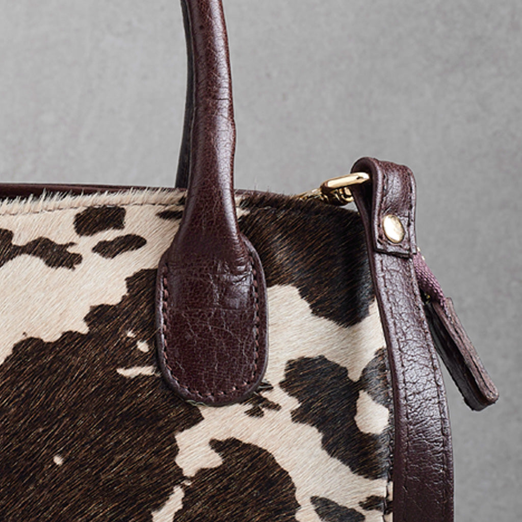 Close-up of a cowhide handbag with dark brown leather handles and trim, featuring a brown, black, and cream cow print pattern and gold-tone hardware.