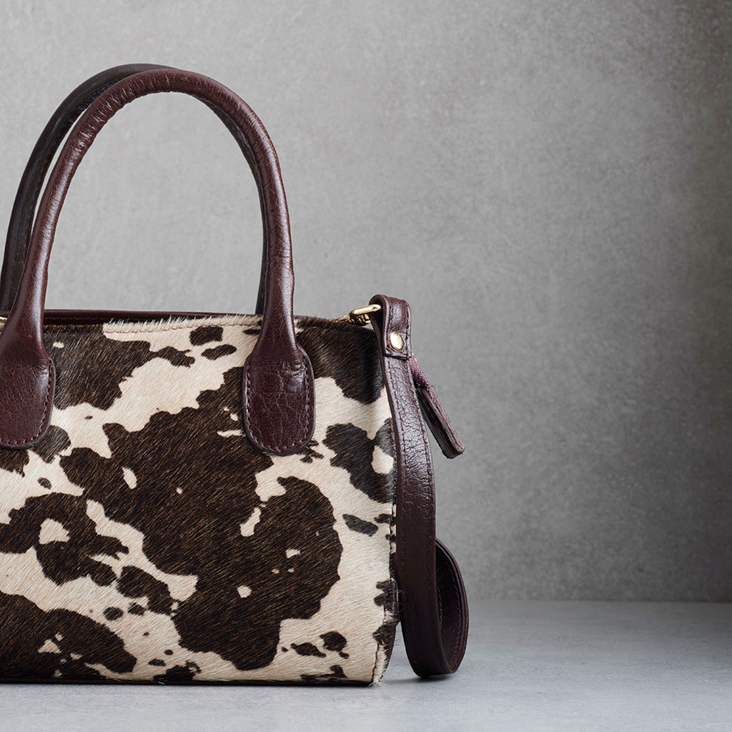 A structured handbag with a brown and cream cowhide pattern and dark brown leather handles and trim, shown against a textured gray background.