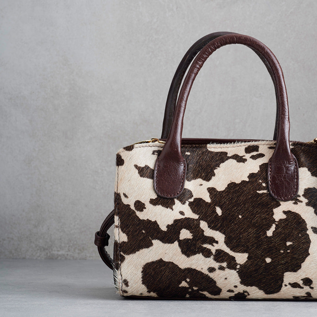 A structured handbag with a brown and cream cowhide pattern body and dark brown leather handles, photographed against a textured gray wall.