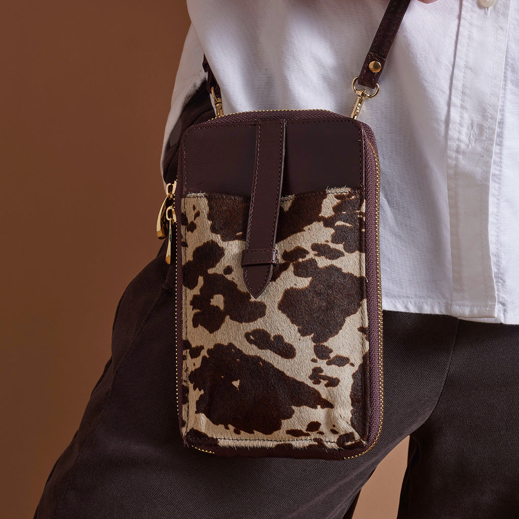 A close-up of a person wearing a brown and beige cow-print crossbody phone pouch bag with dark brown leather trim and a gold zipper, paired with a white shirt and dark trousers.