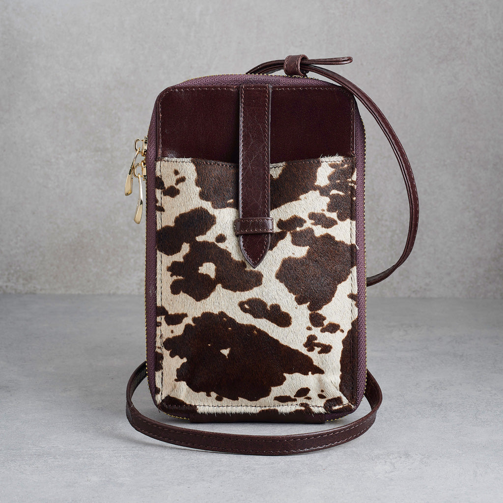 A small crossbody phone bag with a dark brown leather top and strap, and a front pocket featuring a brown and cream cowhide pattern, shown on a gray textured background.