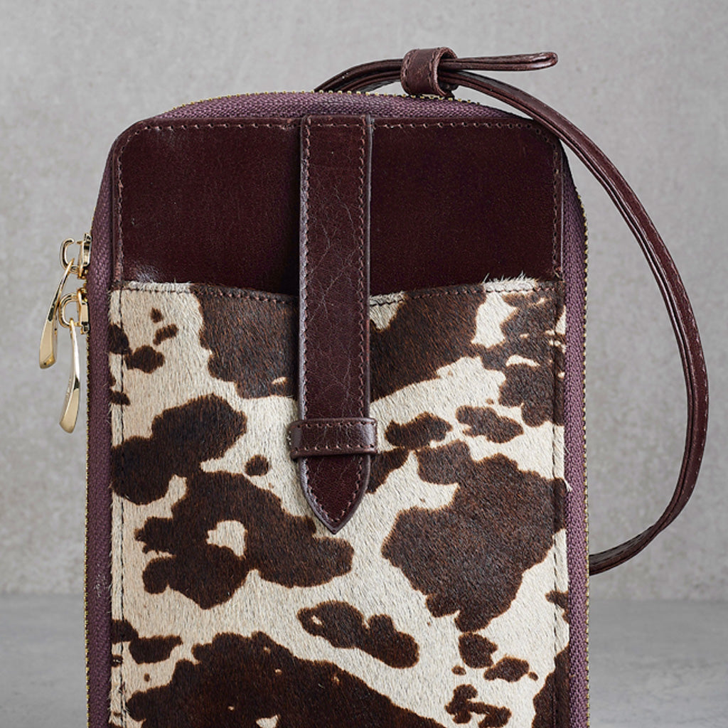 A structured crossbody phone bag featuring a dark brown leather top section and strap, combined with a front pocket made of brown and cream cowhide patterned material, shown against a neutral gray background.