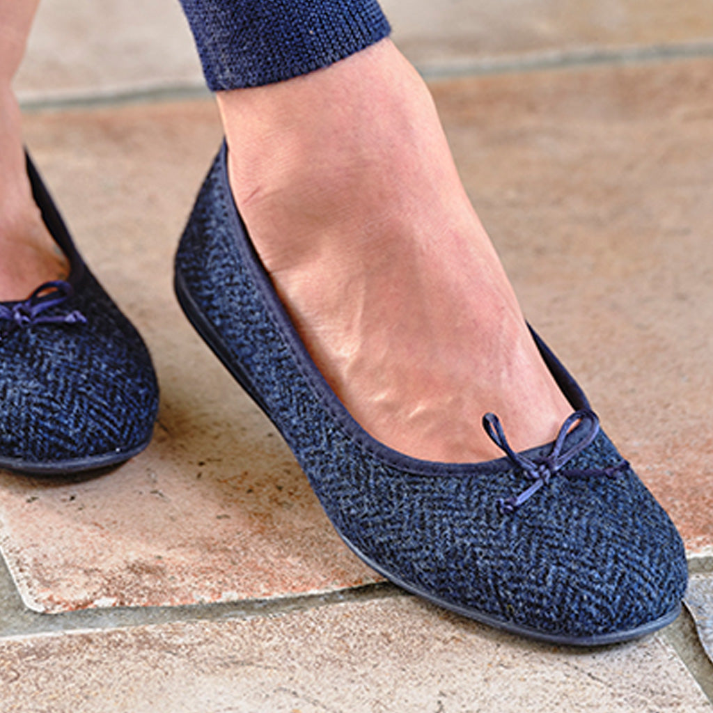 A close-up of a person wearing navy blue herringbone flat shoes with small bow details on the toes, standing on a stone tile floor.