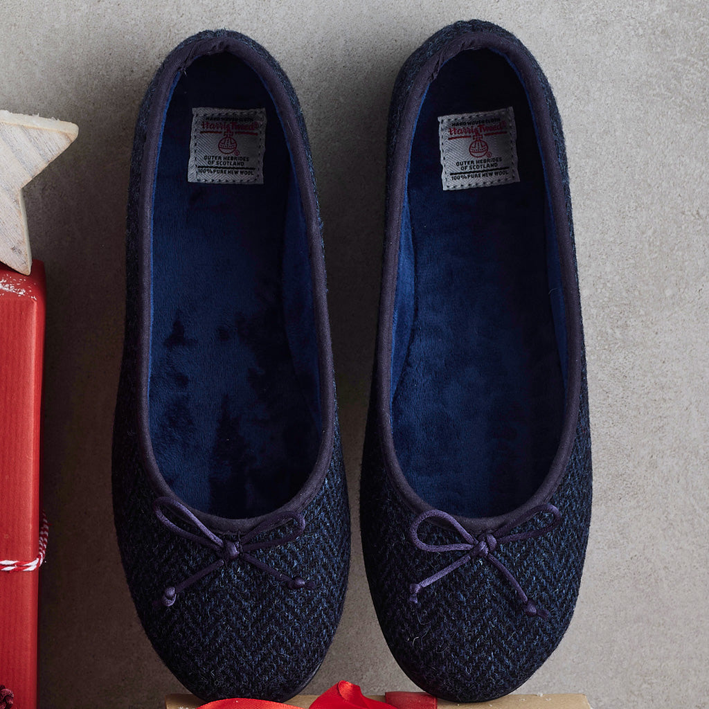 A pair of dark navy blue herringbone wool slip-on slippers with purple fabric trim and matching bow details on the toes, displayed on a neutral gray surface.
