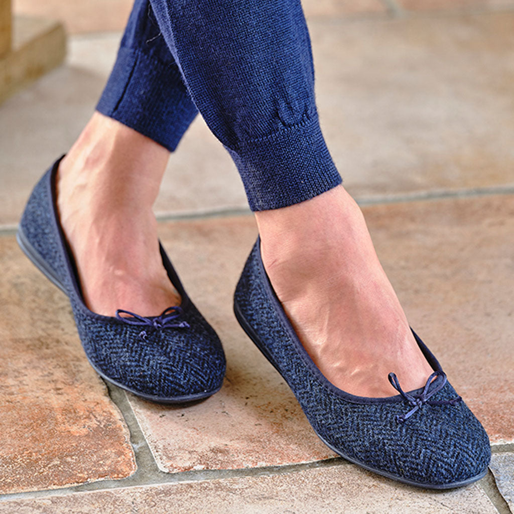 A close-up of a person wearing navy blue textured ballet flats with small bow details, paired with matching navy blue pants, standing on a stone tile floor.